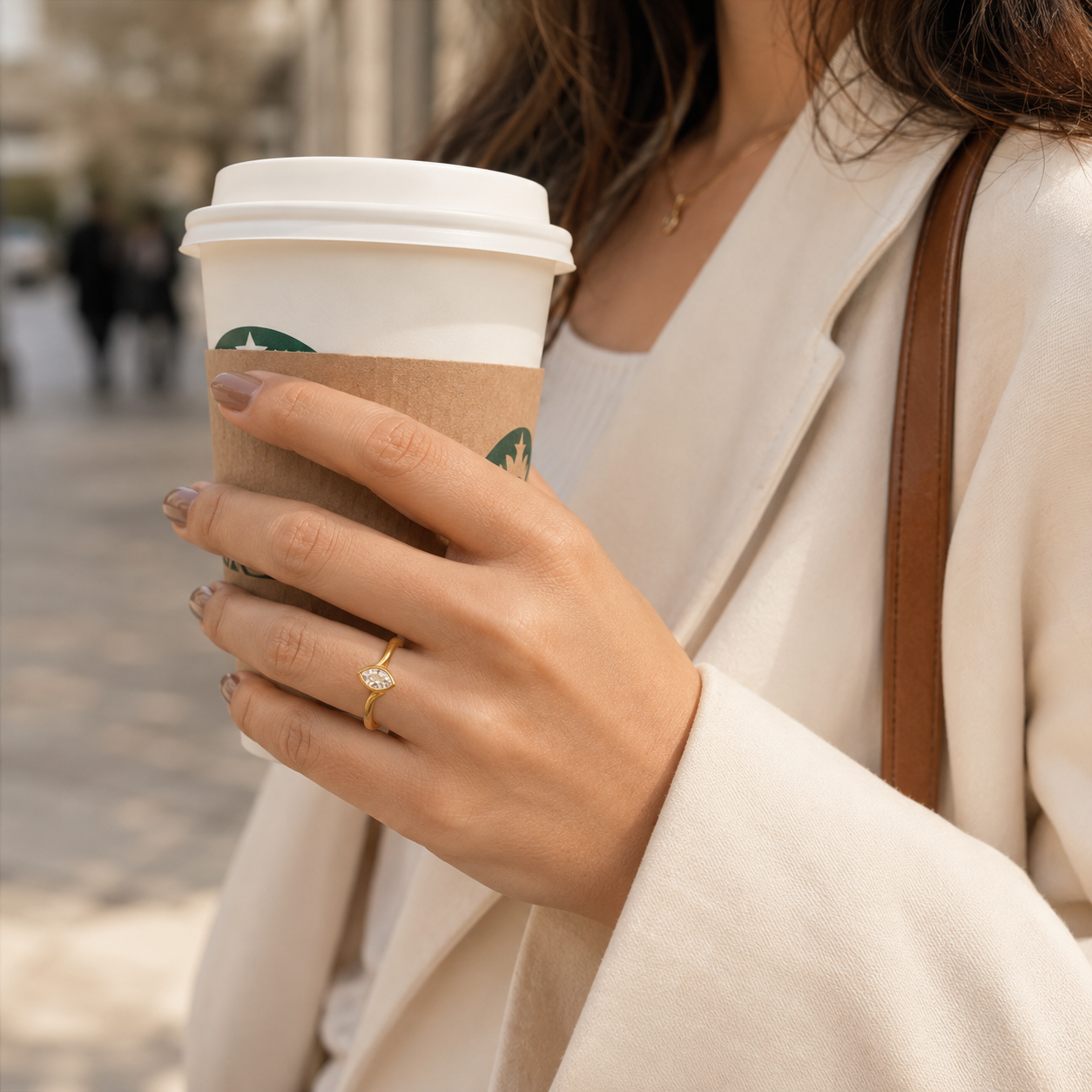 A close-up lifestyle shot of a woman holding a Starbucks coffee cup outdoors while wearing a delicate gold ring with a small clear gemstone on her finger.