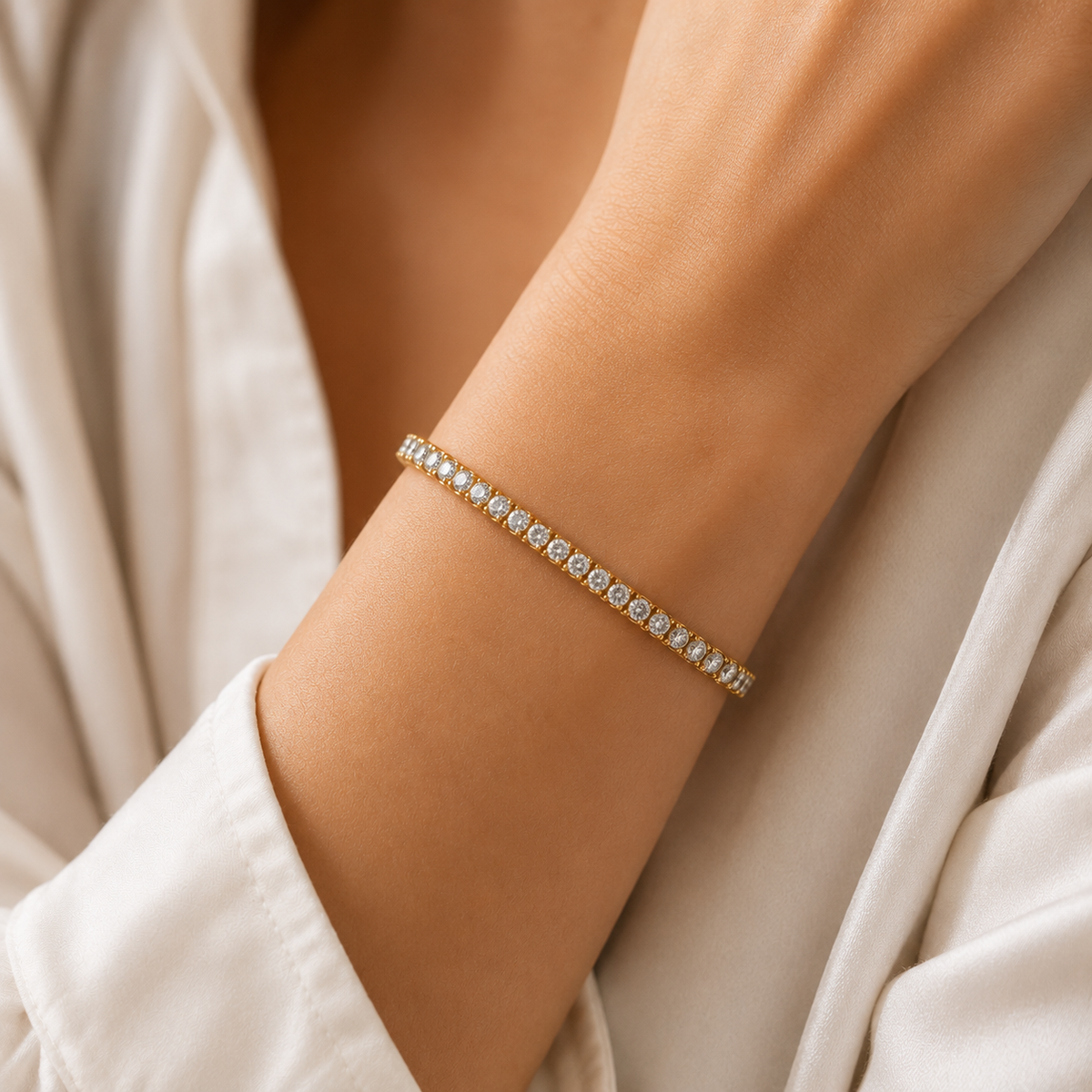 A delicate gold bracelet with a row of small clear gemstones worn on a person's wrist, shown against a soft cream-colored robe.