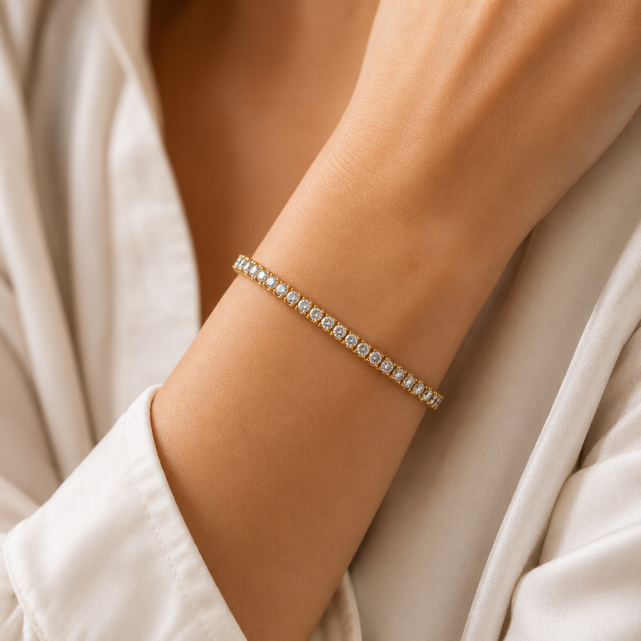 A delicate gold bracelet with a row of small clear gemstones worn on a person's wrist, shown against a soft cream-colored robe.