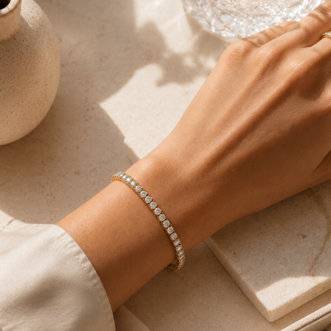 A delicate gold tennis bracelet with small clear stones worn on a woman's wrist, shown in a warm, sunlit lifestyle setting with a textured beige surface, a ceramic vase, and a glass.